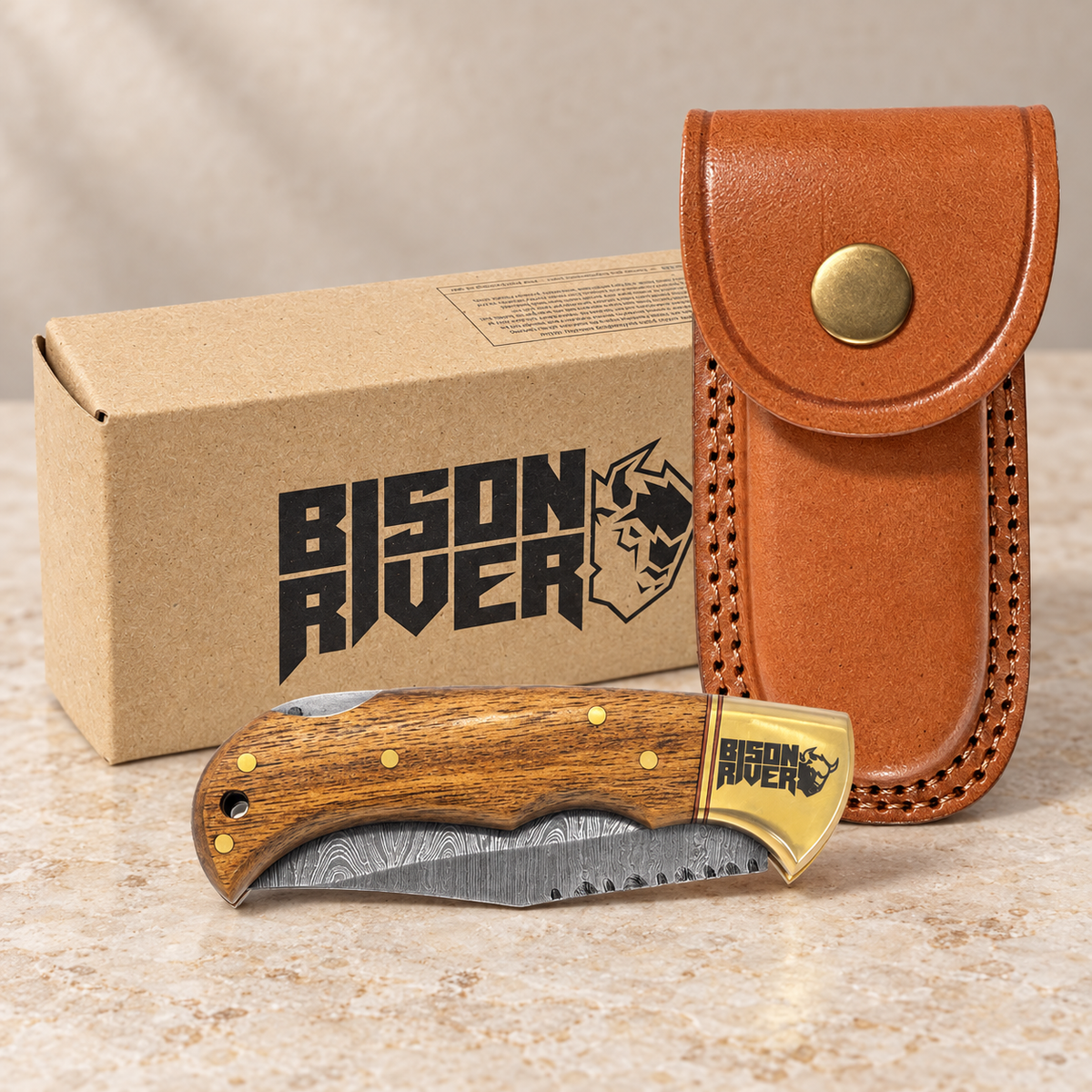 A Bison River folding knife with a wood-grain handle and patterned steel blade, displayed in front of its brown leather sheath and branded cardboard box on a light stone surface.