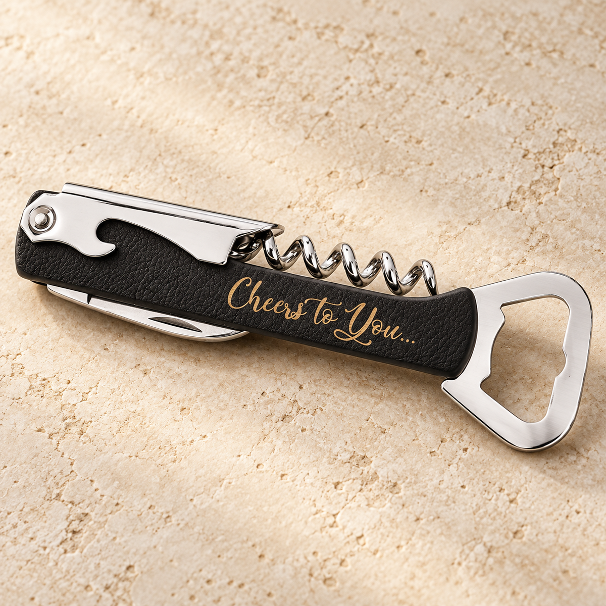 A sleek, black corkscrew wine bottle opener rests on a light, textured surface. The opener features a polished metal corkscrew and a bottle opener, with an engraved message that reads Cheers to You, adding a personal touch.