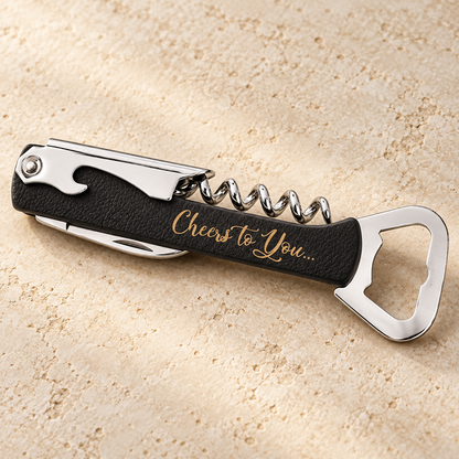 A sleek, black corkscrew wine bottle opener rests on a light, textured surface. The opener features a polished metal corkscrew and a bottle opener, with an engraved message that reads Cheers to You, adding a personal touch.