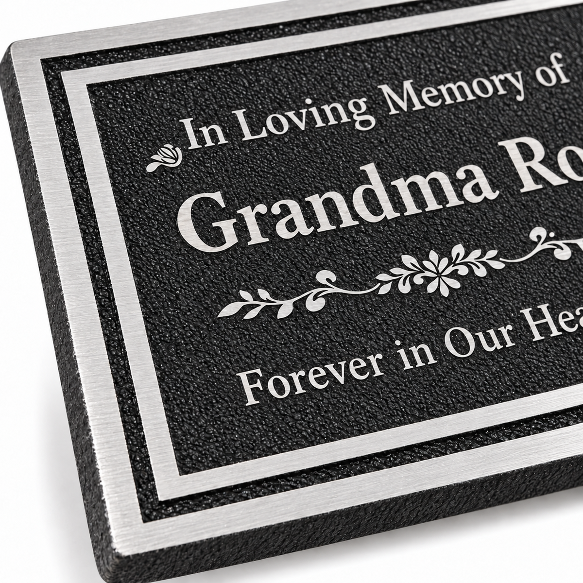 Close-up of a rectangular memorial plaque with a black textured face, silver border, and engraved white text reading 'In Loving Memory of Grandma Rose Forever in Our Hearts.'.