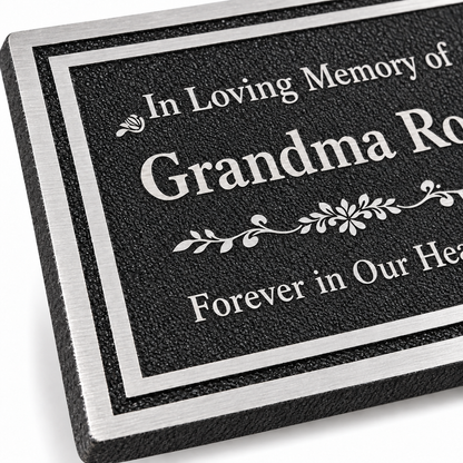 Close-up of a rectangular memorial plaque with a black textured face, silver border, and engraved white text reading 'In Loving Memory of Grandma Rose Forever in Our Hearts.'.