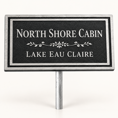 A rectangular black sign with silver border and silver text reading 'NORTH SHORE CABIN' and 'LAKE EAU CLAIRE', mounted on a silver metal stake against a plain white background.