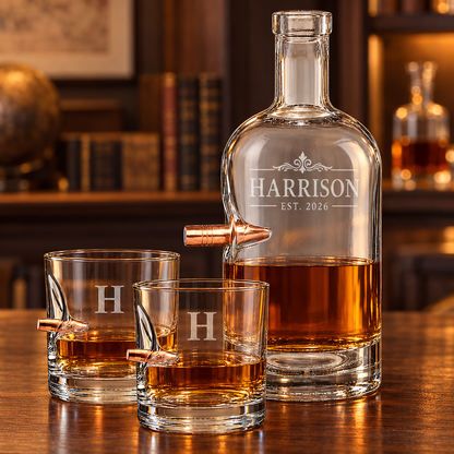A personalized clear glass whiskey decanter bullet embedment and two matching rocks glasses, all engraved with the name HARRISON and EST. 2026, set on a wooden table in a warm bar-like interior.
