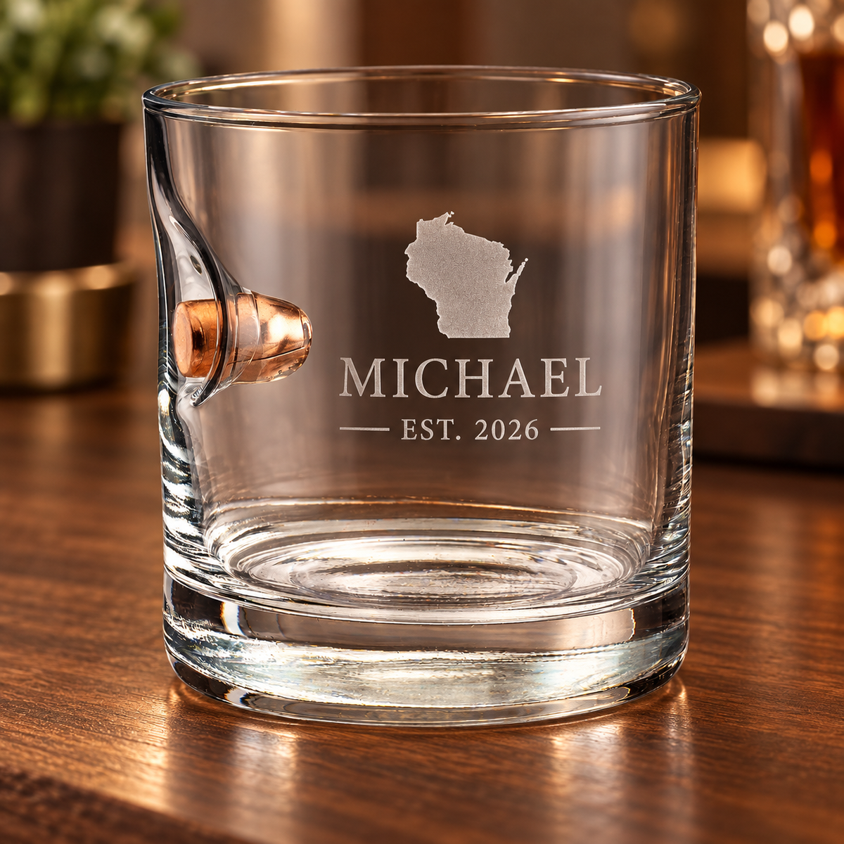 A clear glass rocks tumbler with a handle and a bullet embedment, personalized with an etched Wisconsin state, the name MICHAEL, and EST. 2026, sitting on a wooden bar surface.
