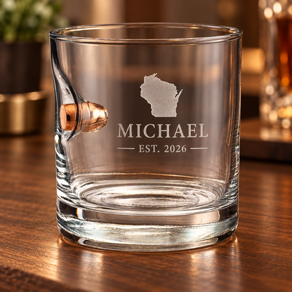 A clear glass rocks tumbler with a handle and a bullet embedment, personalized with an etched Wisconsin state, the name MICHAEL, and EST. 2026, sitting on a wooden bar surface.