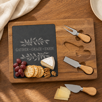 A wooden charcuterie board set on a wooden table, featuring a dark slate insert engraved with 'GATHER GRAZE ENJOY' and leaf motifs, alongside three wooden-handled cheese knives and crackers, grapes, cheese, and a folded cream cloth.