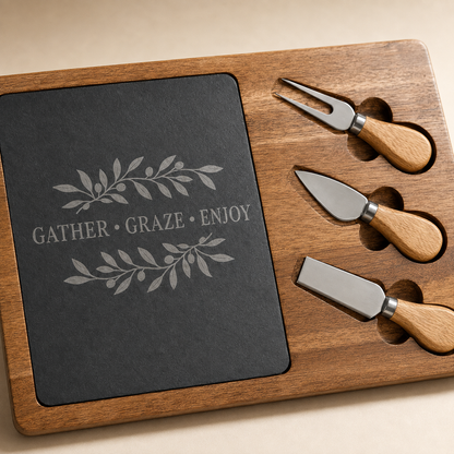 A wooden serving tray set with a dark slate board engraved with 'GATHER • GRAZE • ENJOY' and leaf motifs, plus three metal cheese knives with light wood handles.