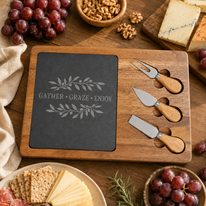 A wooden charcuterie board set on a rustic wooden table, featuring a dark slate insert engraved with 'GATHER • GRAZE • ENJOY' and three wooden-handled cheese knives and a fork.