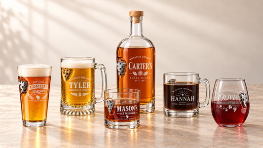 A set of personalized whiskey glasses and a branded bottle of Carter's Small Batch Bourbon Whiskey arranged on a tabletop. The collection includes a pint glass, a beer mug, a lowball glass, a stemless wine glass, and a full-size whiskey bottle, all featuring custom printed labels with names and illustrations.