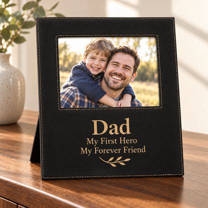 A black leatherette photo frame with gold laser etched lettering that reads 'Dad My First Hero My Forever Friend' and a small leaf motif, standing on a wooden table. The photo insert shows a smiling man carrying a child on his shoulders.