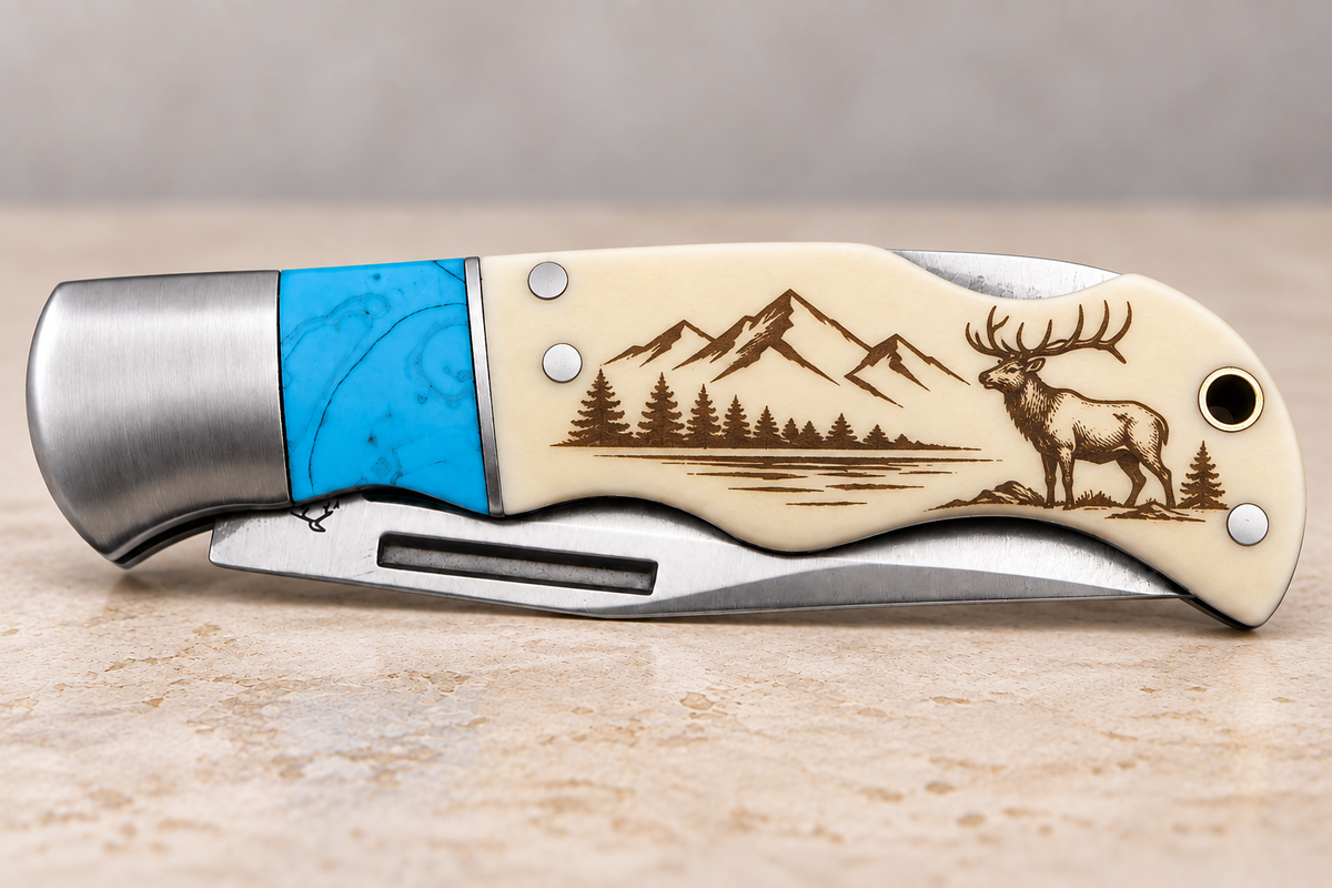 A folding pocket knife with a cream-colored handle featuring a detailed brown engraving of mountains, pine trees, and a deer. The knife has a bright blue inlay on the bolster and a brushed silver metal blade and end cap, resting on a textured beige surface.
