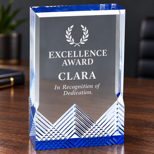 A clear acrylic or glass award plaque with blue patterned edges and base, engraved with the text 'EXCELLENCE AWARD CLARA In Recognition of Dedication' and a laurel wreath, sitting on a wooden desk in an office setting.