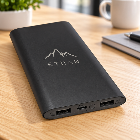 A black portable power bank with a silver mountain logo and the name 'ETHAN' on the front, resting on a light wood desk with a blurred coffee mug and notebook in the background.