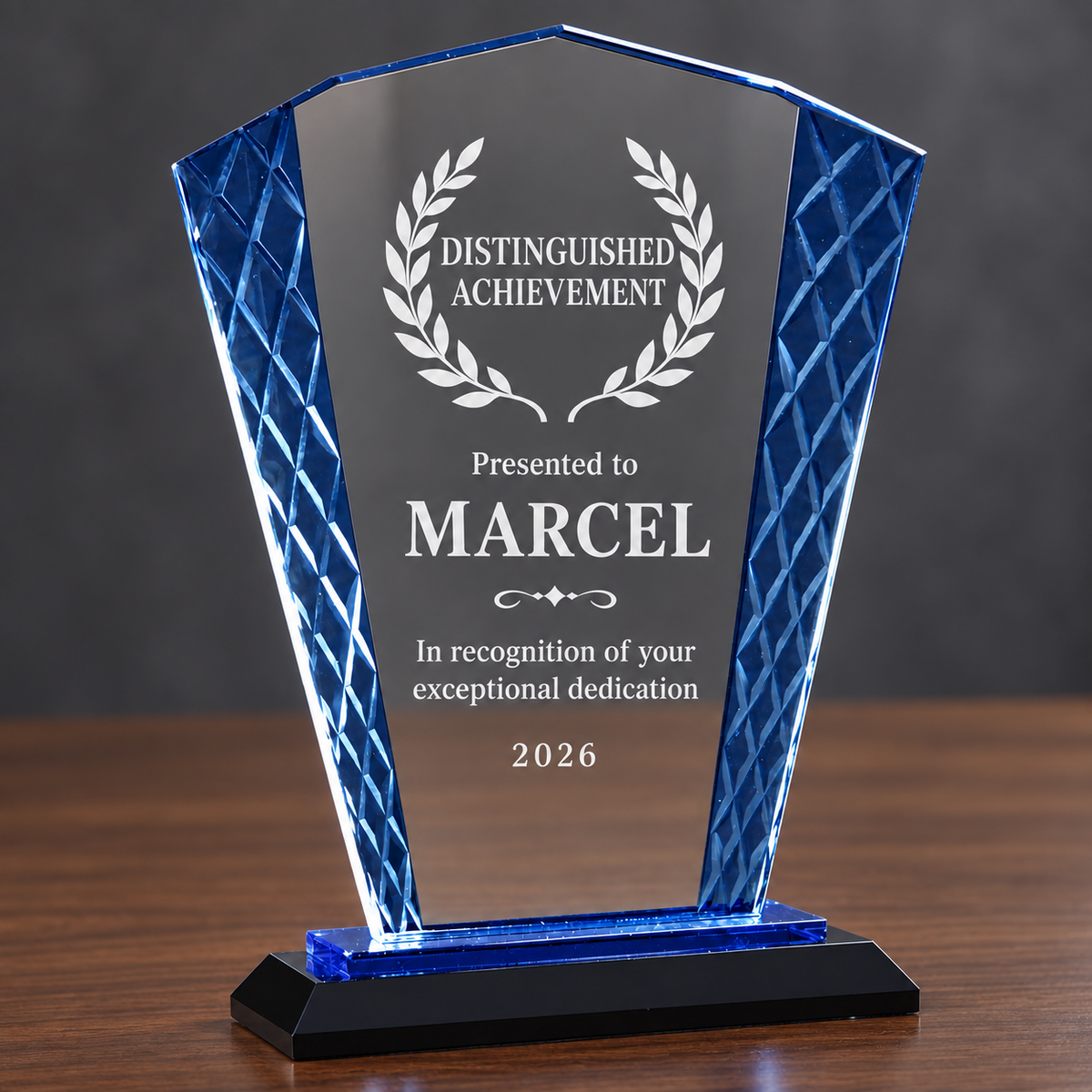 A personalized acrylic award plaque on a black base, featuring a clear central panel engraved with 'Distinguished Achievement' and the name 'MARCEL', flanked by textured blue side panels, set on a wooden surface against a dark gray background.