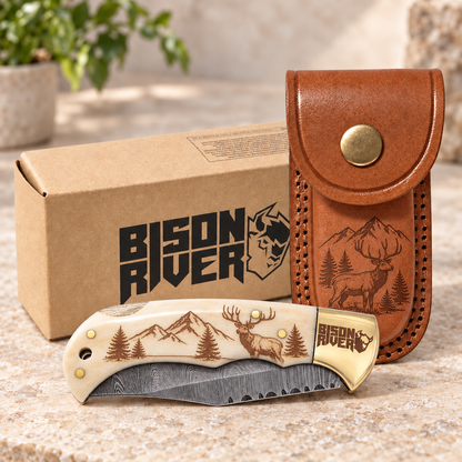 An engraved folding pocket knife with a deer and mountain design on the handle, displayed in front of its brown branded box and a brown leather sheath on a stone surface.