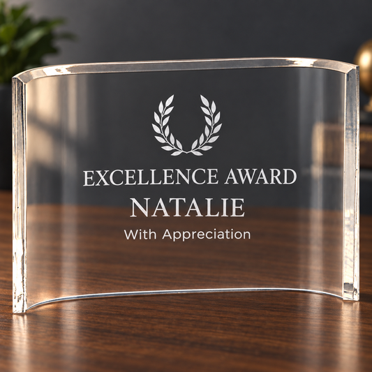 A clear acrylic award plaque standing on a wooden table, engraved with a laurel wreath and the text 'EXCELLENCE AWARD NATALIE With Appreciation'.
