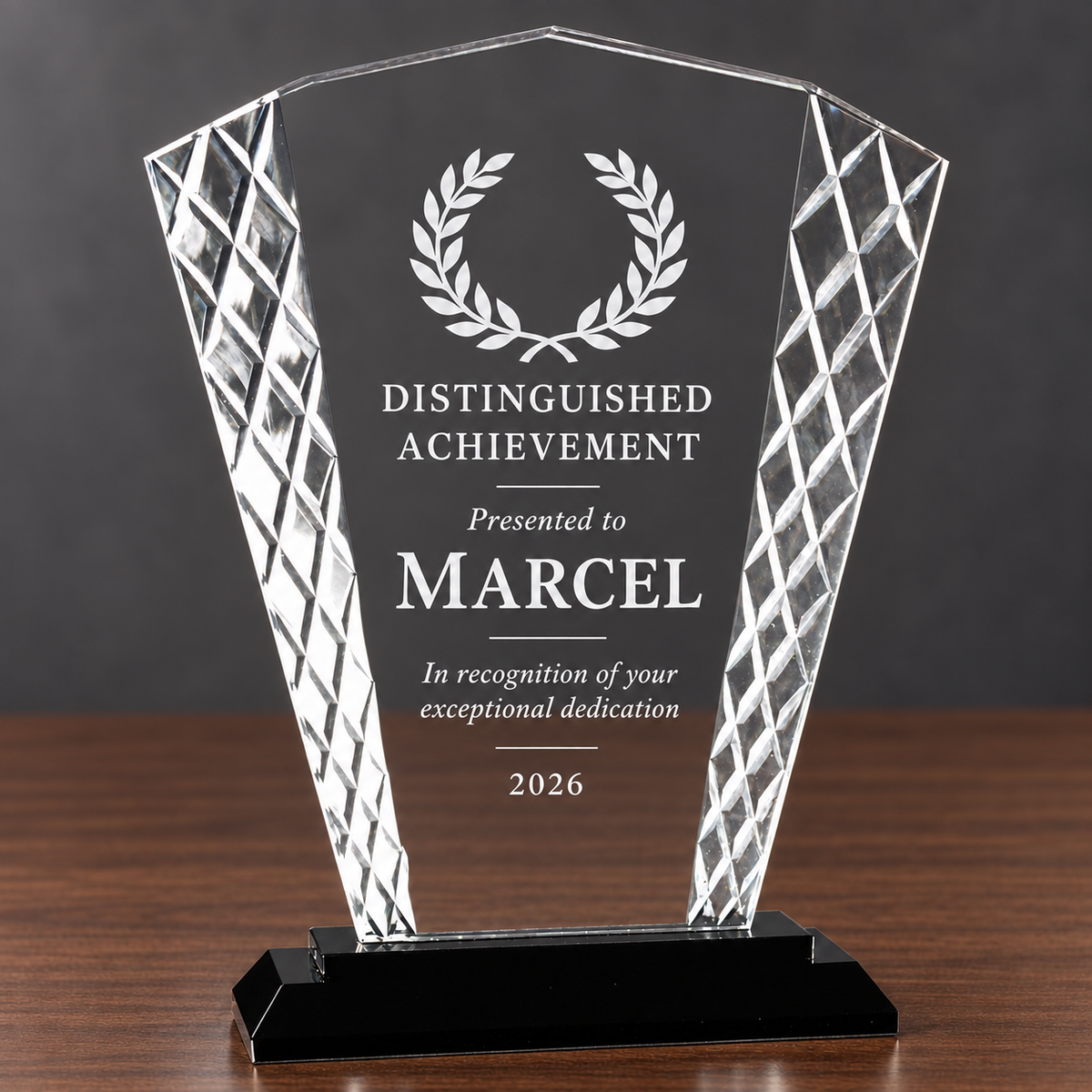 A clear acrylic award trophy with laser-engraved text and a laurel wreath, displayed on a black base over a wooden surface against a dark gray background.