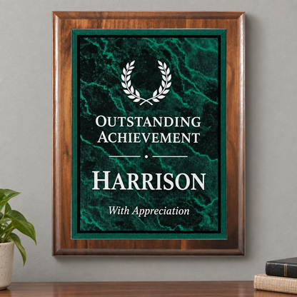 A framed personalized achievement print with a dark green marble background, white laurel wreath graphic, and the text 'OUTSTANDING ACHIEVEMENT HARRISON With Appreciation,' displayed on a wall above a wooden surface with a small plant and books nearby.