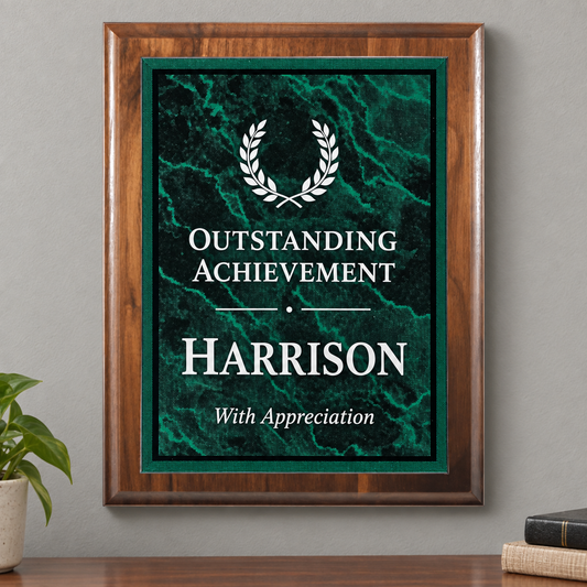 A framed personalized achievement print with a dark green marble background, white laurel wreath graphic, and the text 'OUTSTANDING ACHIEVEMENT HARRISON With Appreciation,' displayed on a wall above a wooden surface with a small plant and books nearby.