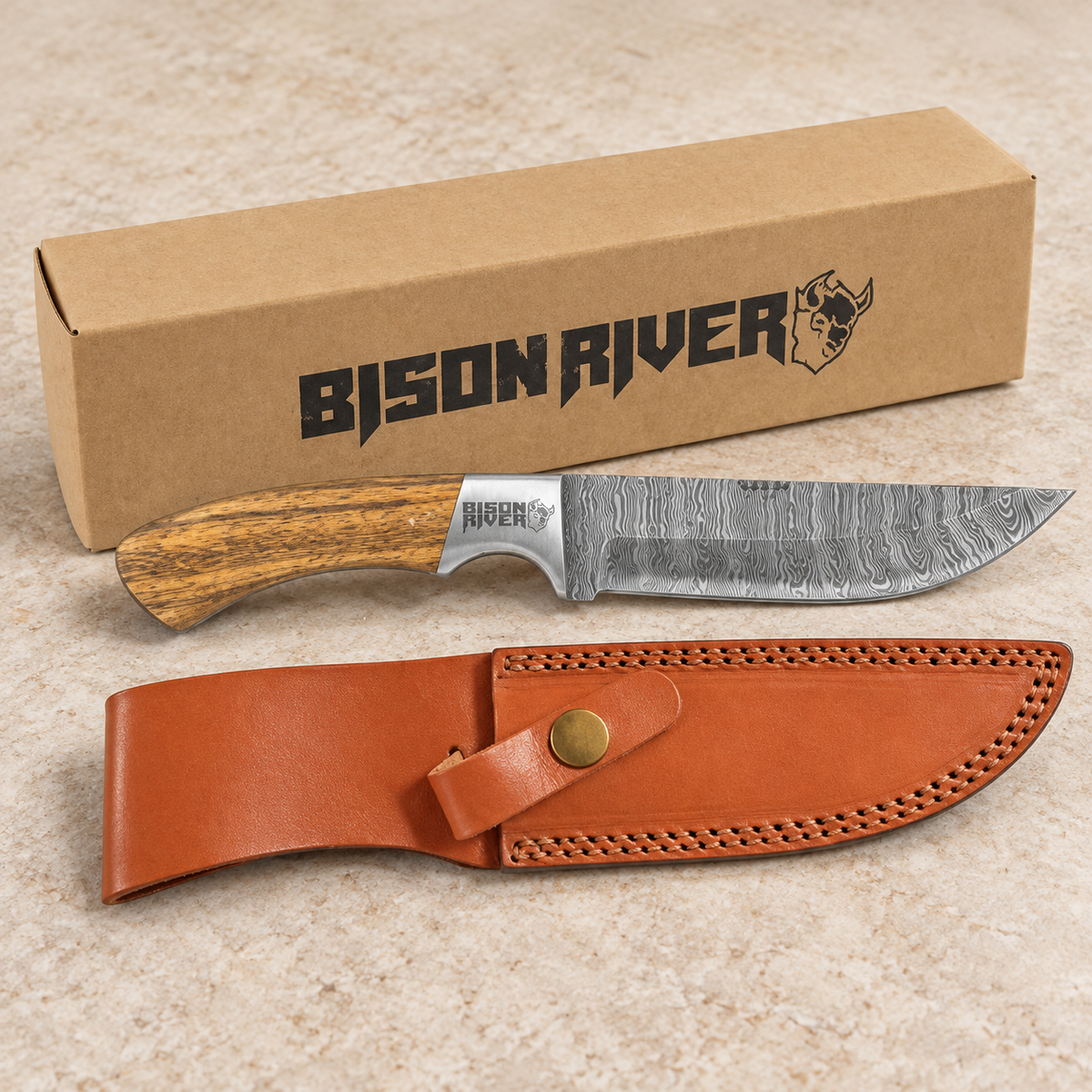 A fixed-blade knife with a Damascus steel blade and wood-grain handle is displayed in front of its brown cardboard box and a brown leather sheath on a light beige stone surface. The box and knife handle both show the Bison River brand.