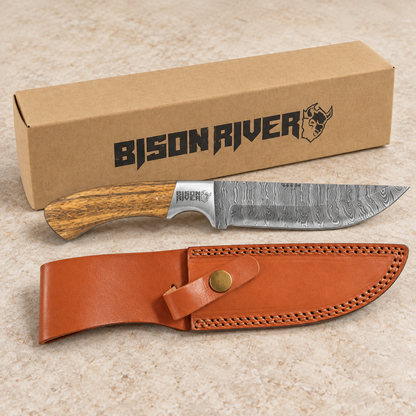 A fixed-blade knife with a Damascus steel blade and wood-grain handle is displayed in front of its brown cardboard box and a brown leather sheath on a light beige stone surface. The box and knife handle both show the Bison River brand.