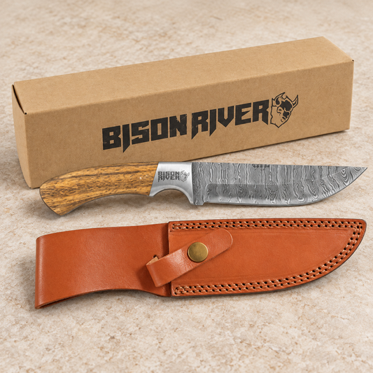 A fixed-blade knife with a Damascus steel blade and wood-grain handle is displayed in front of its brown cardboard box and a brown leather sheath on a light beige stone surface. The box and knife handle both show the Bison River brand.