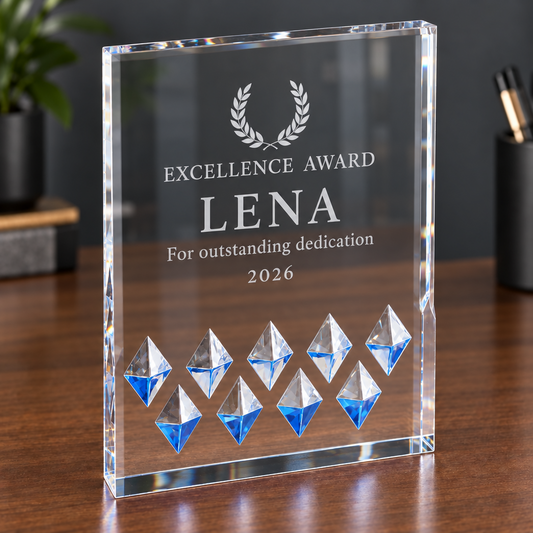 A clear acrylic award plaque standing on a wooden desk, engraved with the text 'EXCELLENCE AWARD LENA For outstanding dedication 2026' and a laurel wreath above the name. The base has geometric crystal-like cutouts with blue and silver facets.