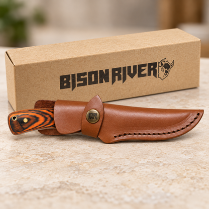 A Bison River fixed-blade knife with a striped wood handle and a brown leather sheath, displayed in front of its branded brown cardboard box on a light stone surface.
