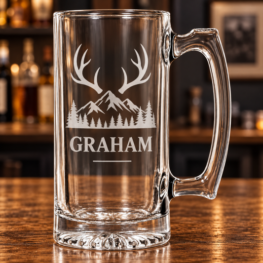 A clear glass beer mug with a handle, featuring a white etched design of antlers, mountains, pine trees, and the name 'GRAHAM' on a wooden bar counter with a blurred bar background.