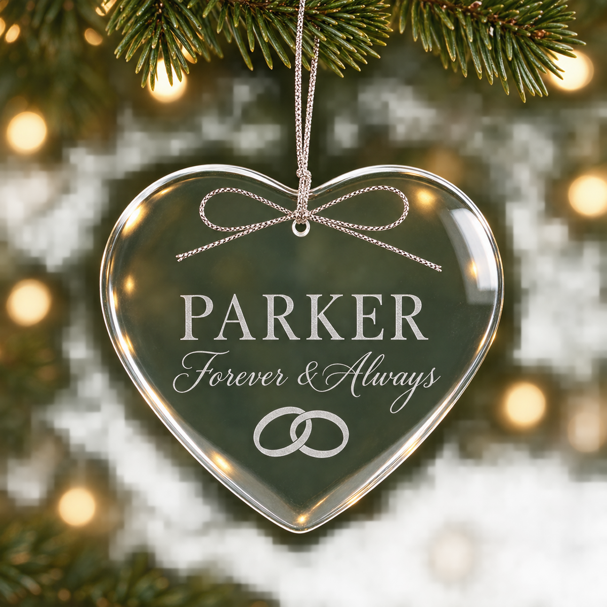 A heart-shaped clear acrylic Christmas ornament hanging from a pine branch, engraved with the name 'PARKER', the words 'Forever & Always', and two interlocking rings, against a blurred festive background with warm lights.