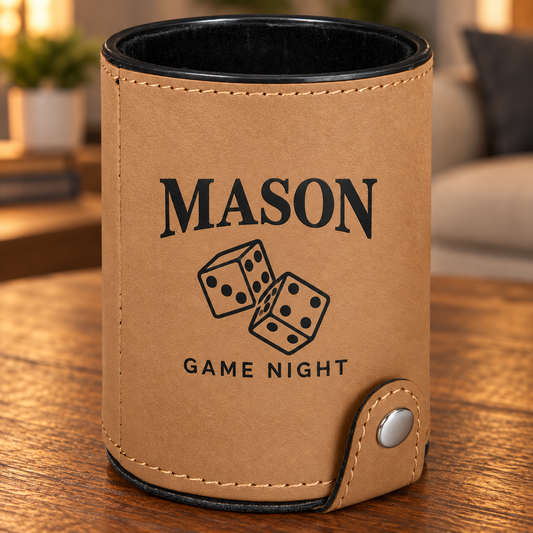 A personalized tan leather dice cup with black trim, featuring the text 'MASON GAME NIGHT' and a graphic of two dice. The cup is shown resting on a wooden table in a warm, blurred living room setting.