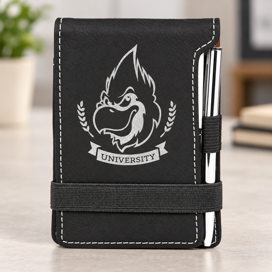 Black leatherette mini notepad with silver UNIVERSITY sample engraving, white stitching, elastic strap and silver pen on a light desk.