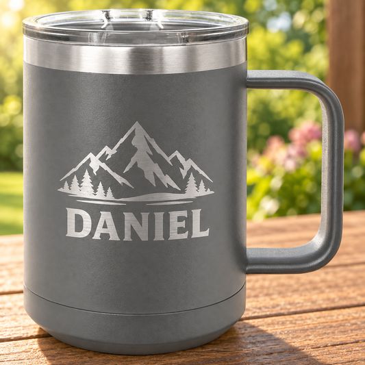 Gray 15 oz coffee mug with silver DANIEL and mountain engraving, a matching handle and clear lid, on a wooden table.