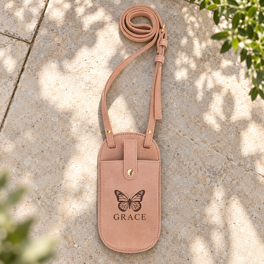 Blush pink Creekside leatherette phone pouch with GRACE and a butterfly design, a matching thin shoulder strap and gold-tone snap, on a stone tabletop.