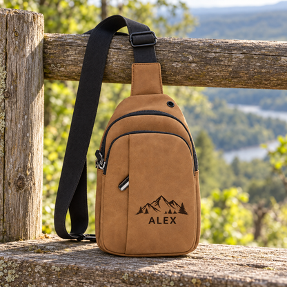 Brown Creekside Explorer leatherette crossbody bag engraved with ALEX and a mountain design, with a black adjustable strap, at a wooded overlook.