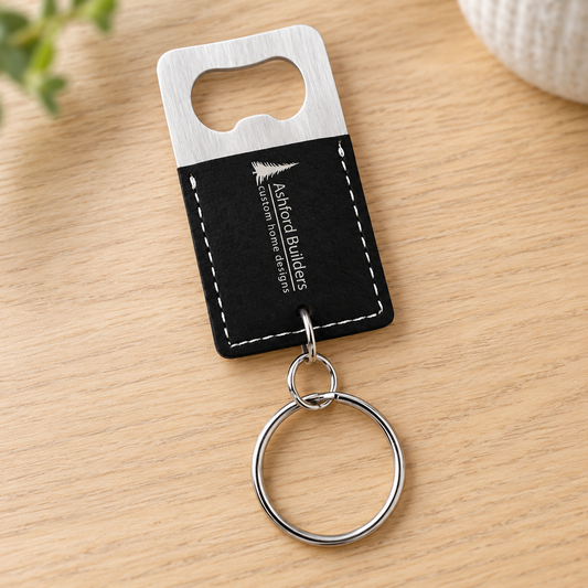 Black and silver rectangular bottle-opener keychain with Ashford Builders sample engraving and bottom-mounted rings on a pale wood surface.