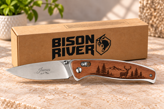 A personalized folding pocket knife with a wooden handle engraved with a deer and forest, displayed in front of its brown cardboard packaging box on a stone surface.