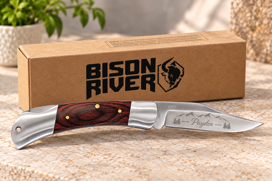 A custom pocket knife with a reddish-brown wood handle and a silver steel blade engraved with 'Payden' and mountain graphics, displayed in front of its brown cardboard packaging box with the 'Bison River' logo.